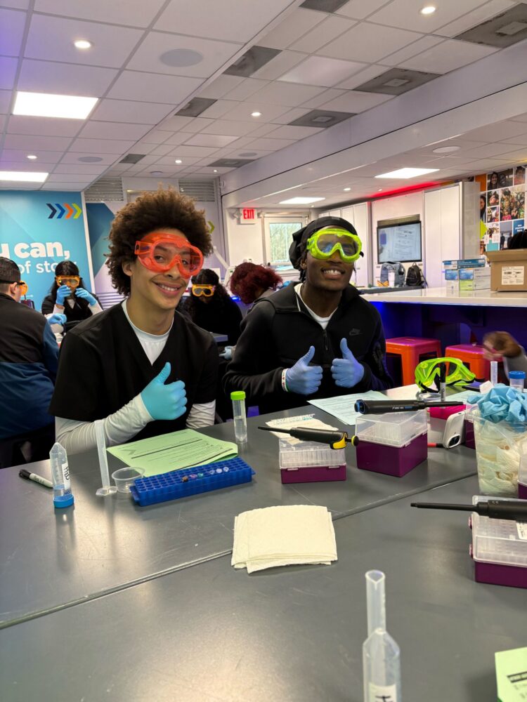 two students wearing bright colored goggles work in a laboratory setting.